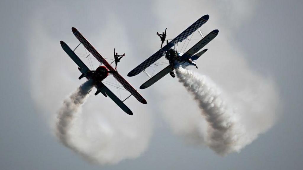 Two planes flying in the sky with smoke trails coming off the back. On the top of each plane is an acrobat.