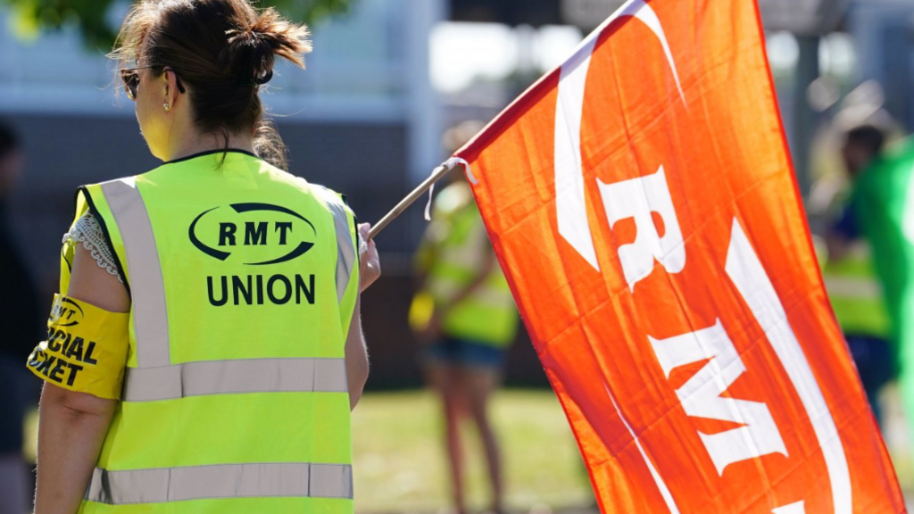 This picture shows a person participating in an RMT (National Union of Rail, Maritime and Transport Workers) union picket or protest. The individual is wearing a high-visibility vest labeled "RMT UNION," holding an orange flag with "RMT" on it, and has an armband that says "OFFICIAL PICKET."