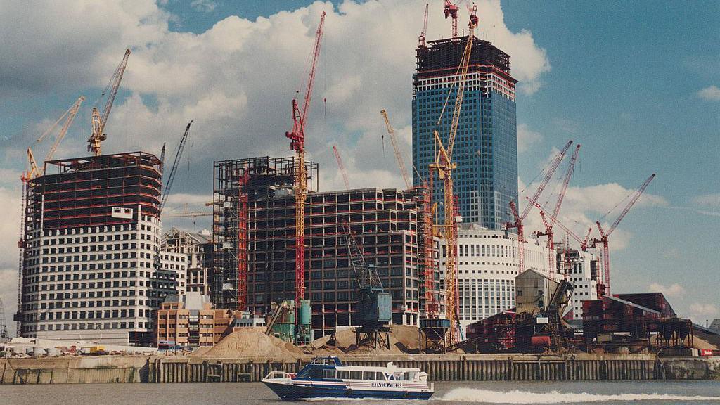 The docks from the water, showing skyscrapers being built. There are lots of cranes and a boat on the river