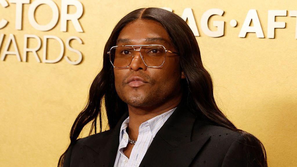 Law Roach wearing a black suit and silver framed glasses photographed by a yellow backdrop at the actors awards