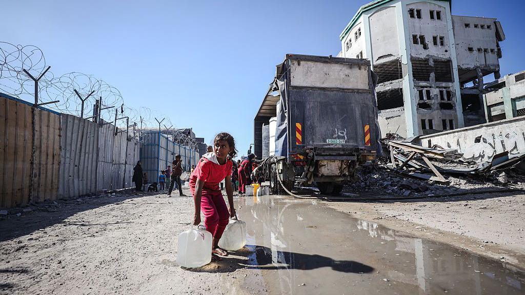 A young girl in a pink top and trousers carries to large bottles of water on the side of the road