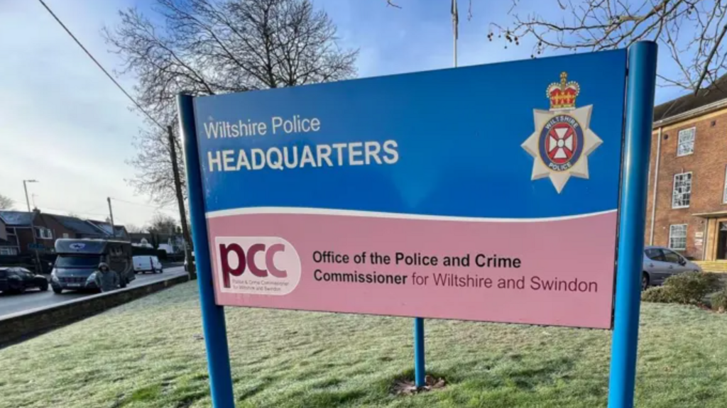 A sign for Wiltshire Police set onto grass with the police headquarters in the background. A road can be seen to the left of the sign.