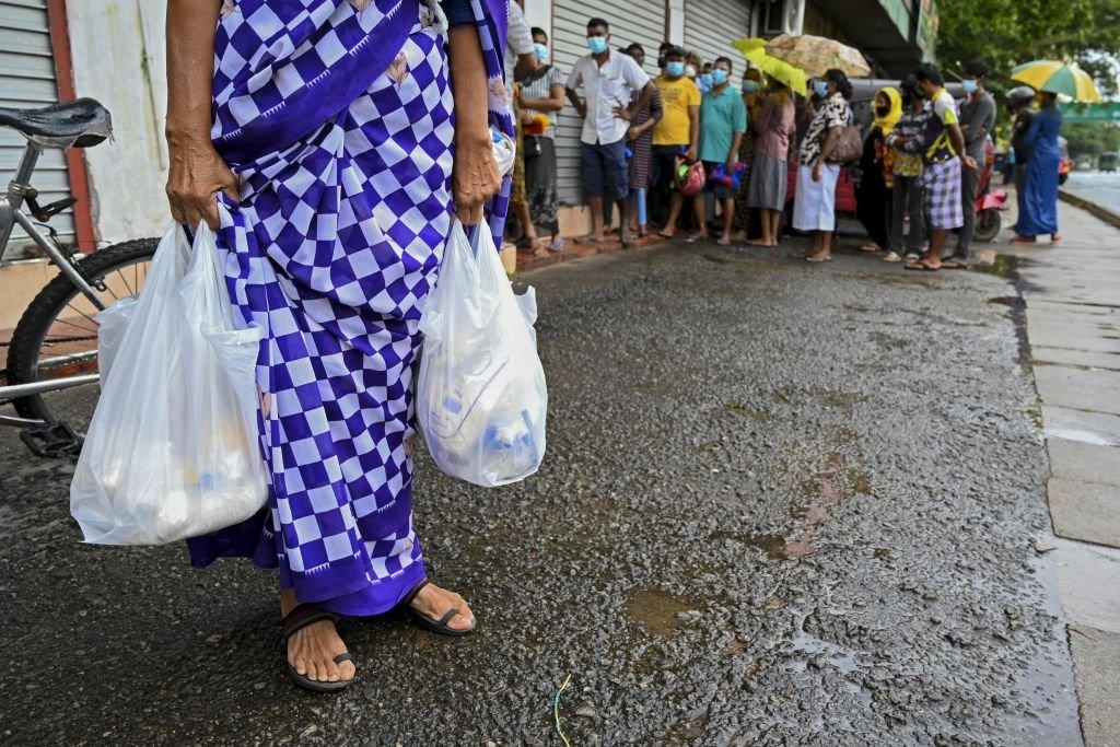 Woman carrying food bags in Colombo while people wait in queue outside a state-run supermarket