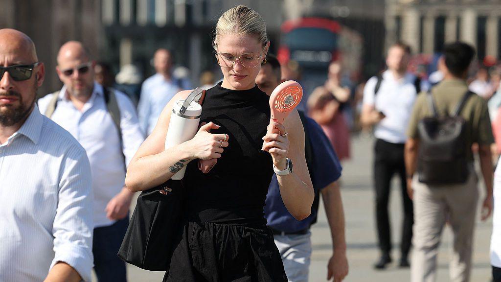 A woman in a black dress uses a personal fan as she walks in the sunshine across London Bridge during a heatwave in central London.