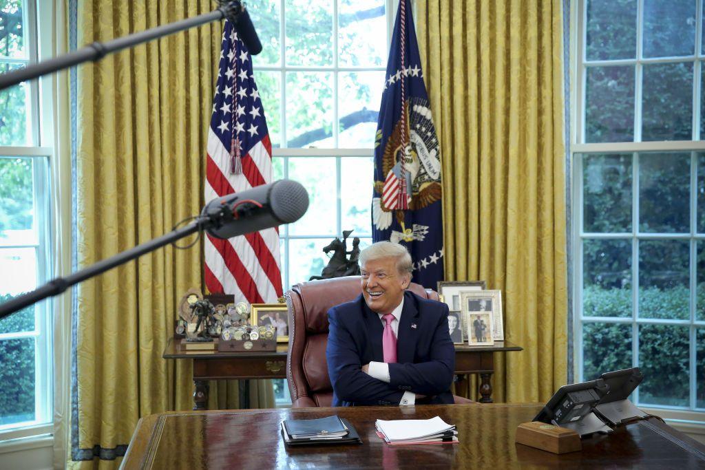 A smiling Trump sits at his Oval Office desk in the White House in front of cameras and TV crews.