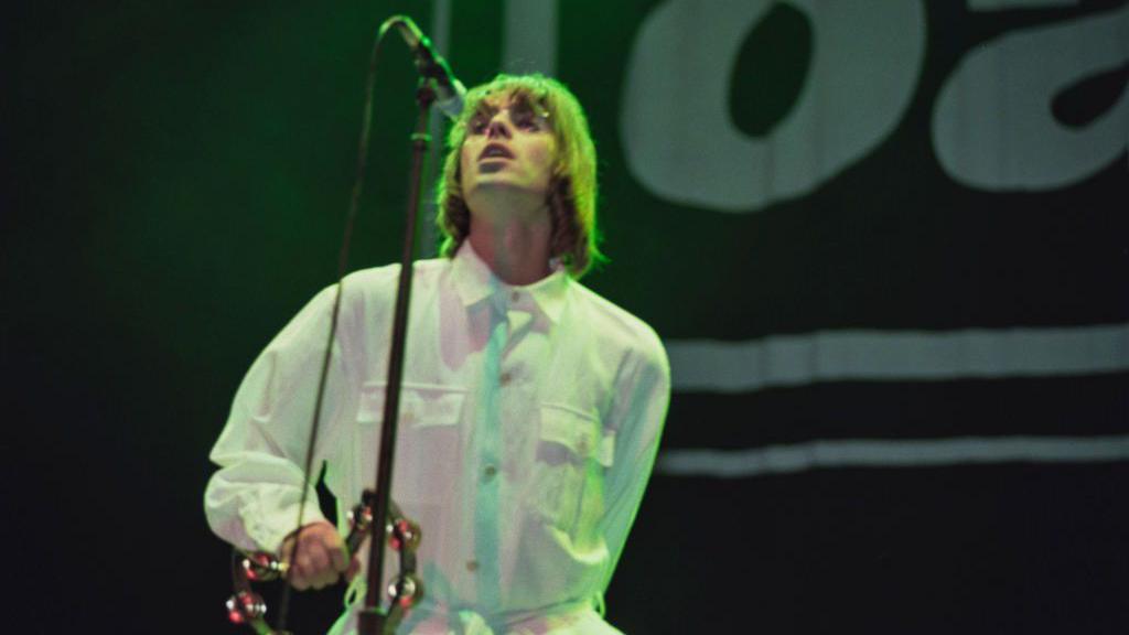 Liam Gallagher on stage at Knebworth, wearing a white shirt and holding his microphone stand