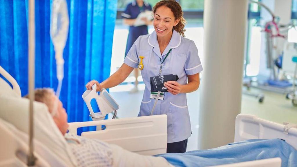 A stock image showing a smiling, female nurse at a patient's hospital bedside