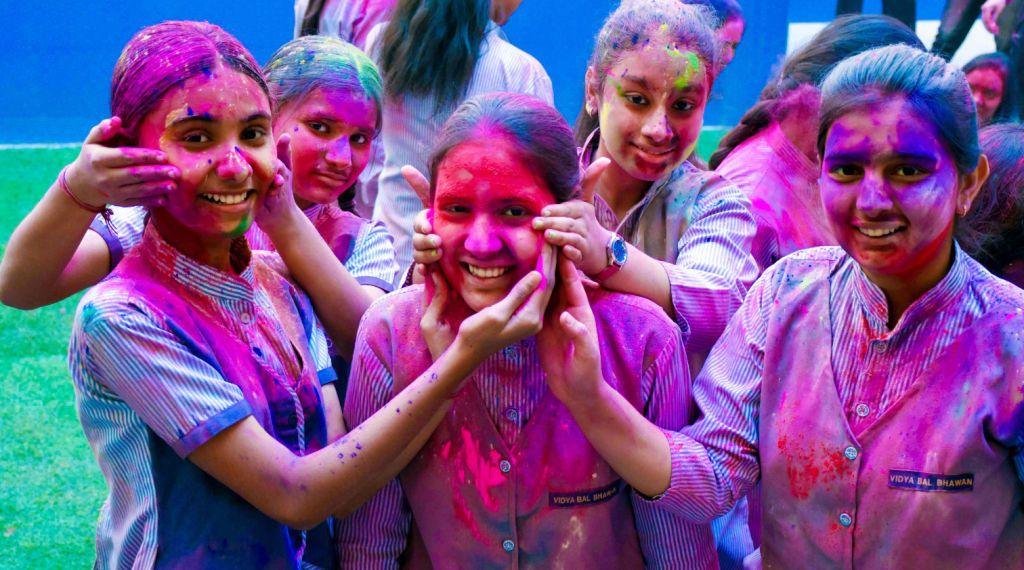 A group of girls in school uniform wearing striped shirts and waistcoats smile while covering one another's faces in brightly coloured paint