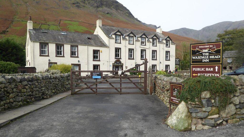 Exterior of the Wasdale Head Inn. It is painted cream and is surrounded by hills. The building is shaped like two large houses. There is a sign showing the pub's name outside. Its gate is closed and it is surrounded by a wall made of stones.