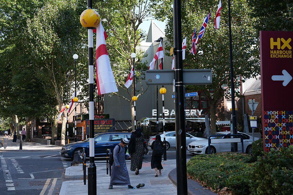 St George's flags and Union Jack flags hang from lampposts and a zebra crossing light on a street in London.