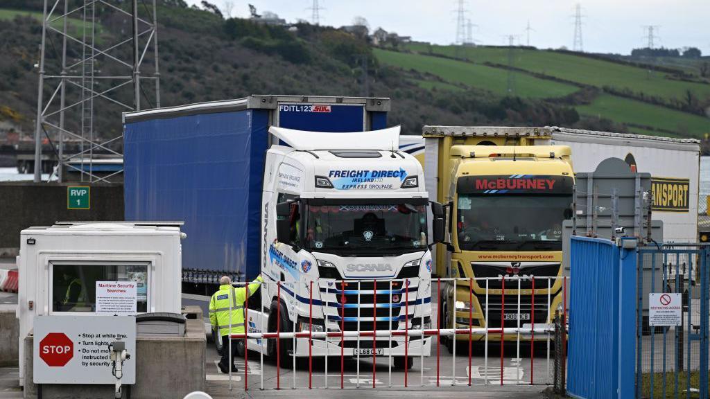 A man dressed in a high-visibility jacket has his hand reached to gather documents from a truck driver.