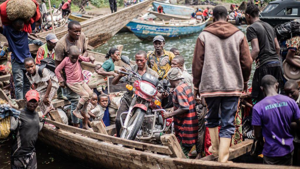 Displaced Congolese people flee the area of Minova by boat. The small, wooden boat is packed and one man has even loaded his motorbike onto the vessel.