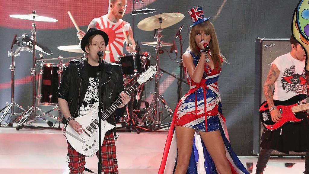 Patrick Stump from Fall Out Boy and Taylor Swift stand on a stage at the 2013 Victoria's Secret fashion show. Patrick (left) wears a black fedora, a black ts-shirt, a black leather jacket, red tartan trousers and holds a white guitar. Taylor Swift (right) wears a dress with a union jack on it with a matching cape and top hat. Her microphone is red.