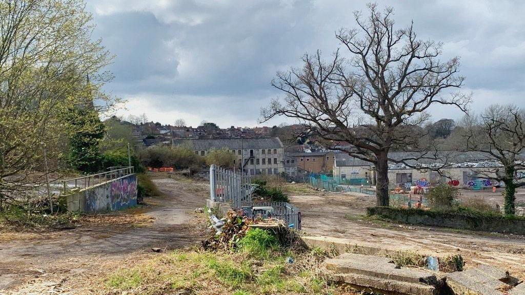 The Saxonvale site in Frome in early 2021. There are several derelict buildings in the background that have graffiti on them, with scrubland in the foreground.