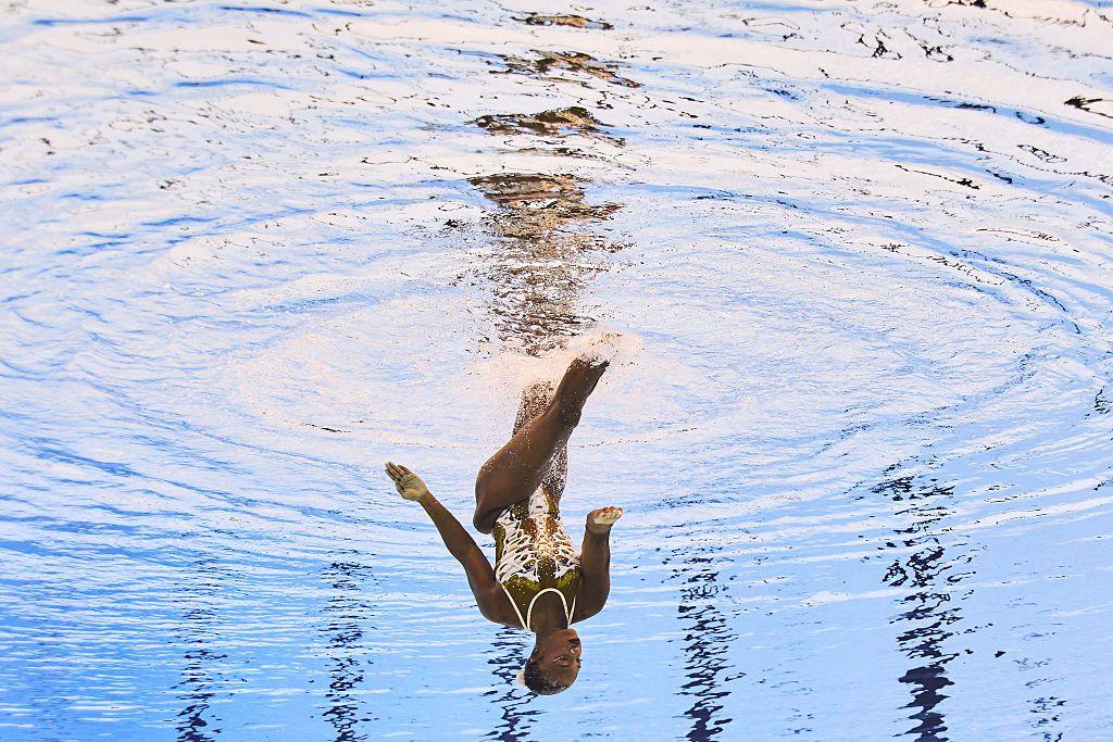 A woman wearing a white-and-gold swimming costume and matching which hairband swims upside-down in an artistic manner. She is competing in an event called the Women's Solo Free.