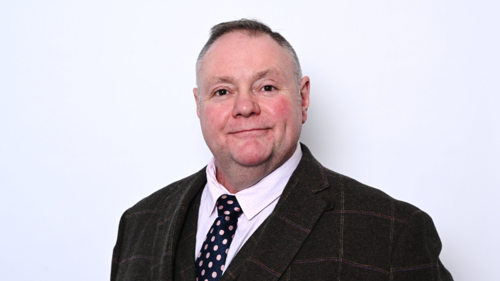 A man with short dark hair in a dark suit with a dark blue patterned tie in front of a white background