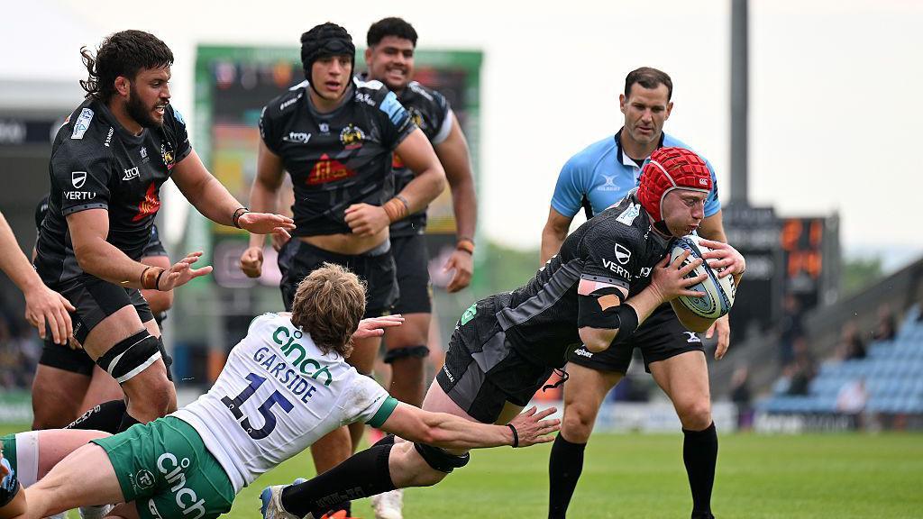 A rugby player in black wearing a red scrum cap dives over the line despite the attempts of a tackler wearing the number 15 to stop him from doing so. The referee and other players look on.