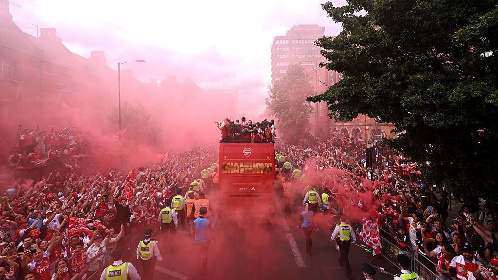 Wide shot of the Arsenal FC victory bus parading down the streets, police officers in yellow hi-vi jackets flanking the vehicle. On both sides of the bus are large crowds of fan, red flare smoke billowing in the air