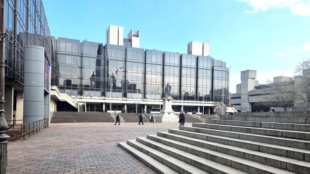 Portsmouth civic offices in Guildhall Square. It is a modern-looking building which wraps around two sides of the square and is faced with black glass. In the foreground are the white stone steps which lead up to the Guildhall. In the centre of the square is a large black statue of Queen Victoria on a stone plinth.