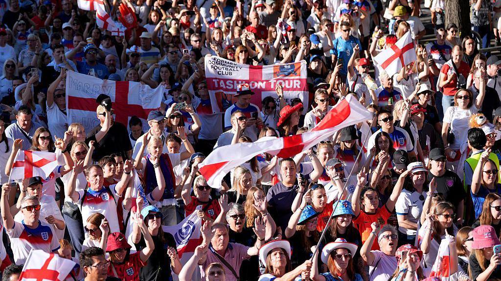 England fans taking part in a fan walk to the stadium prior to the UEFA Women's EURO 2025 Semi-Final match between England and Italy. The tightly packed crowds of people are mostly wearing England football shirts and some are wearing England hats and waving large England flags.
