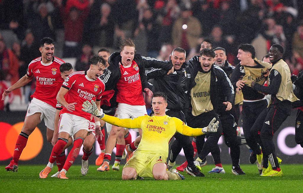 Benfica goalkeeper Anatoliy Trubin slides on his knees with arms wide in celebration as teammates and substitutes in red sprint toward him, leaping and shouting in the rain under floodlights, after he scored a dramatic fourth goal to send his side through to the Champions League play-off round with victory over Real Madrid.