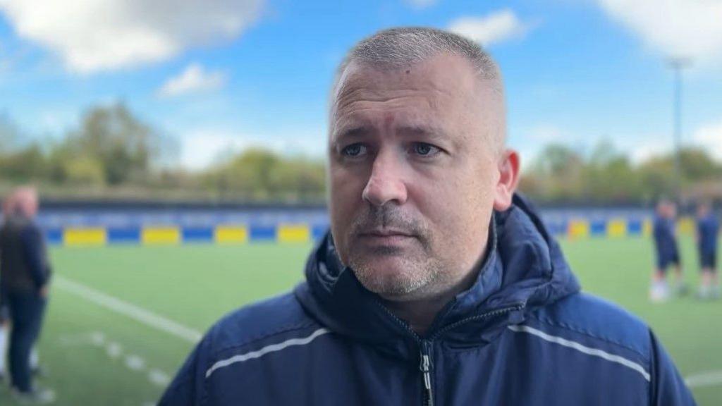 Hereford FC manager Paul Caddis is standing at the side of a football pitch. He is wearing a navy blue coat and is looking towards the left hand side of the shot.