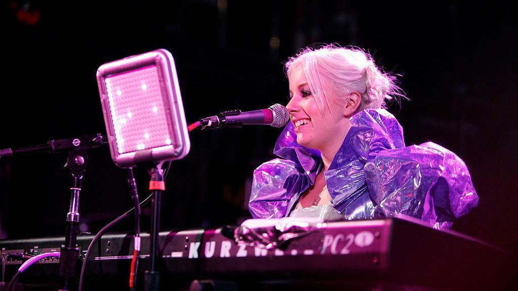 Victoria Mason as a younger artist performing on stage. She is sitting behind a keyboard, wearing a purple shiny costume with puffy sleeves and an exaggerated collar. She is singing into a microphone and smiling, with her bleach blonde hair pulled back into a messy bun, with smaller pieces pulled out at the front. The image is dark with a pink and purple hue.