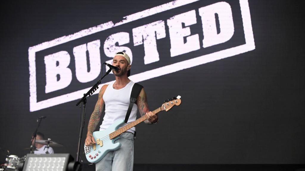 A member of the band Busted sings and plays bass guitar on stage, with a large “BUSTED” sign displayed behind him.