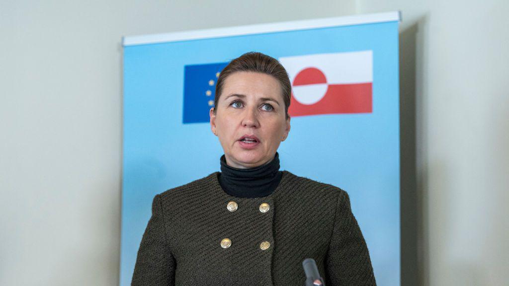 Denmark’s Prime Minister Mette Frederiksen, wearing a dark green textured blazer with gold buttons and a black turtleneck, speaks at a press event. She stands in front of a light blue backdrop featuring the flags of Greenland and the European Union. Her expression is serious as she addresses the audience, with a microphone visible in the foreground.