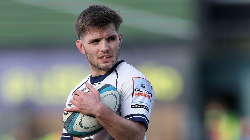 Harry Randall holds the ball close to his chest with his right arm while standing on the pitch
