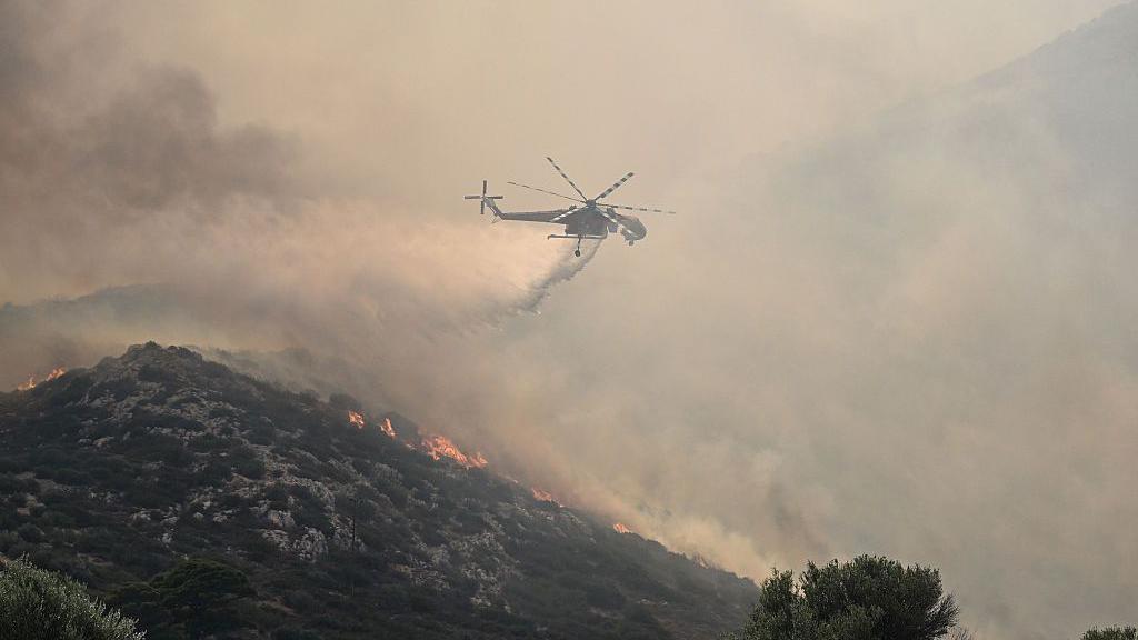 A helicopter drops water over vegetation that is smoking and on fire