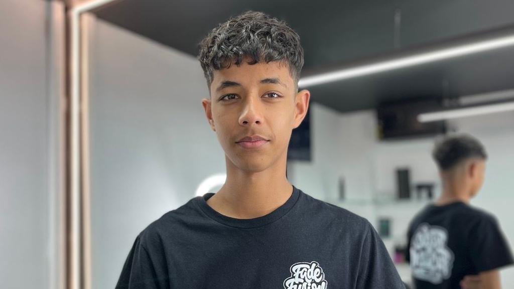 A boy is wearing a black t-shirt and has brown curly hair. He is standing in a barber shop with white walls and a black ceiling.