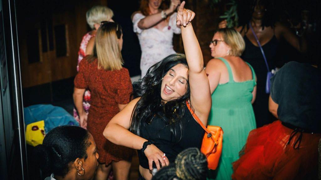 A woman with long dark hair is dancing with her hand raised in the air. She is surrounded by other women, some of whom are also dancing.