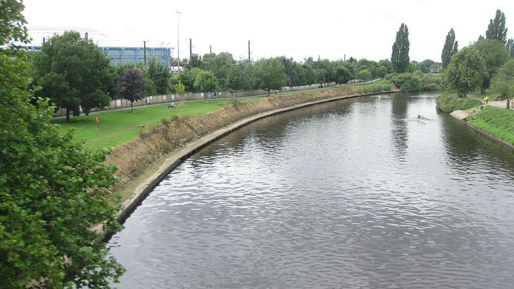 A picture of a river with footpaths on either side. To the left, trees can be seen lining the footpath with a fence running along the left.