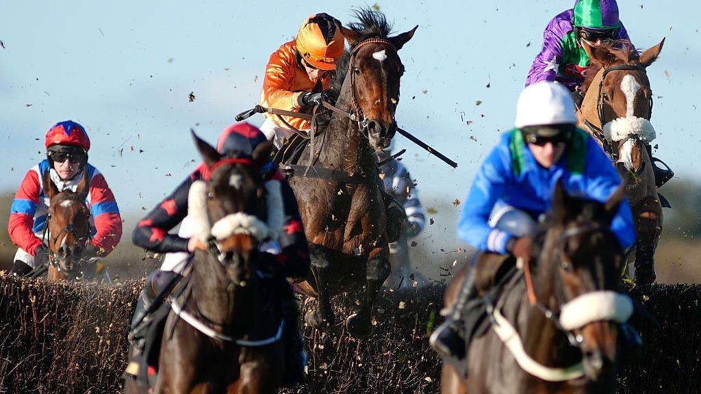 Jockeys and their horses jump over a hurdle, with debris flying around. The jockeys are wearing colourful uniform.
