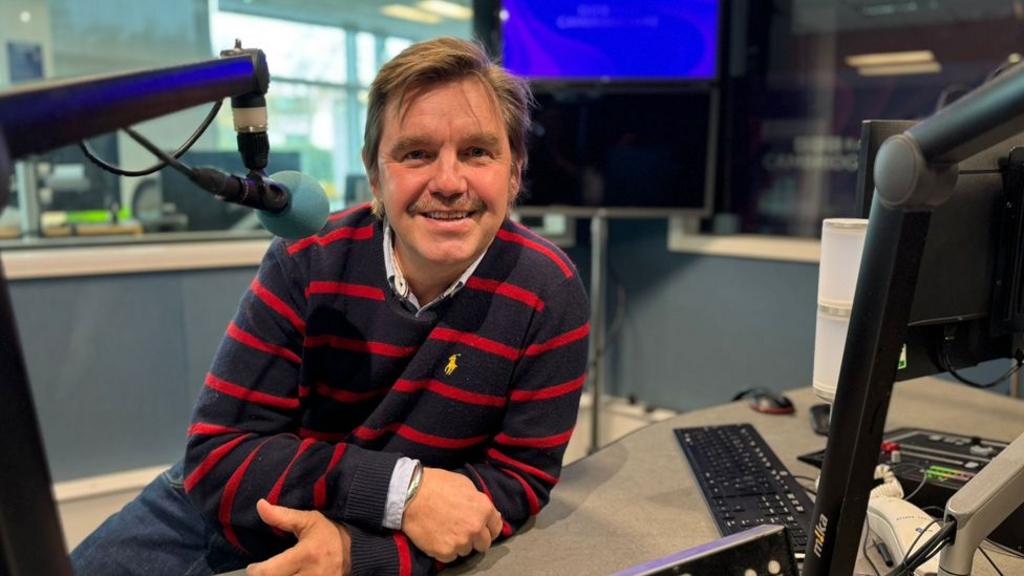 Nik Johnson sitting at the desk in a BBC Radio Cambridgeshire studio, wearing a red and navy blue striped jumper