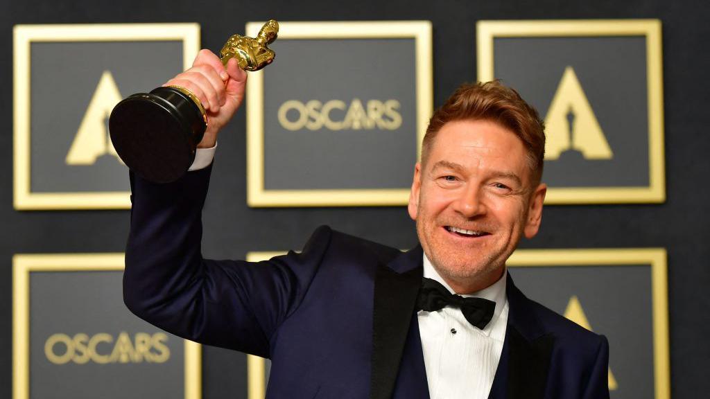 A man with strawberry blonde hair and stubble wearing a dark blue tuxedo with a black bow tie, smiling and holding up a gold Oscars trophy in the air. Behind him is a black and gold wall with Oscars branding on it.