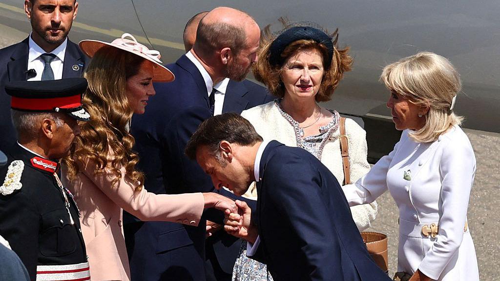 On an airport tarmac Emmanuel Macron kisses the Princess of Wales's hand, while a step behind them, Prince William speaks to Brigitte Macron.