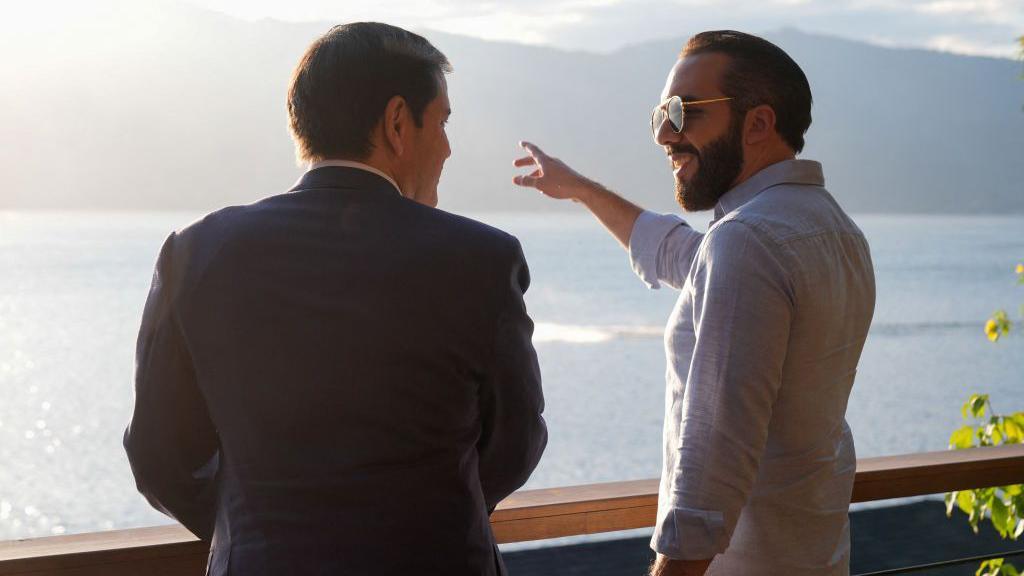 US Secretary of State Marco Rubio in a dark blue suit looks at El Salvador's President Nayib Bukele, who is wearing a light blue shirt and sporting dark sunglasses. The two men are overlooking the lake outside President Bukele's residence