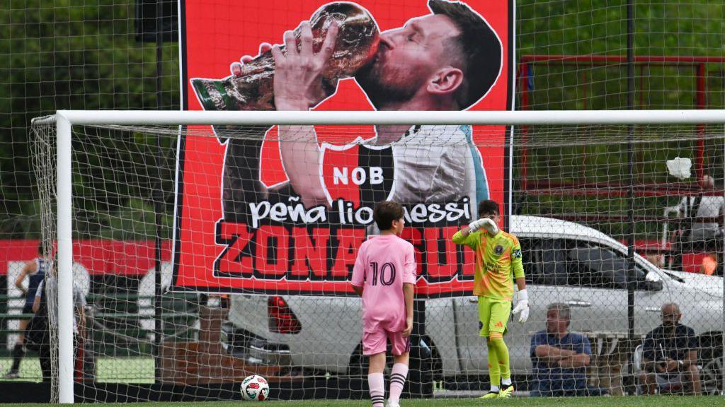 Thiago Messi stands on a football pitch in front of a banner of his father, Lionel Messi, kissing the World Cup