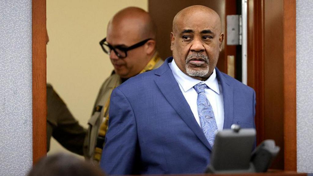 Duane Davis, a bald black man in his 60s, enters a courtroom while wearing a blue suit.