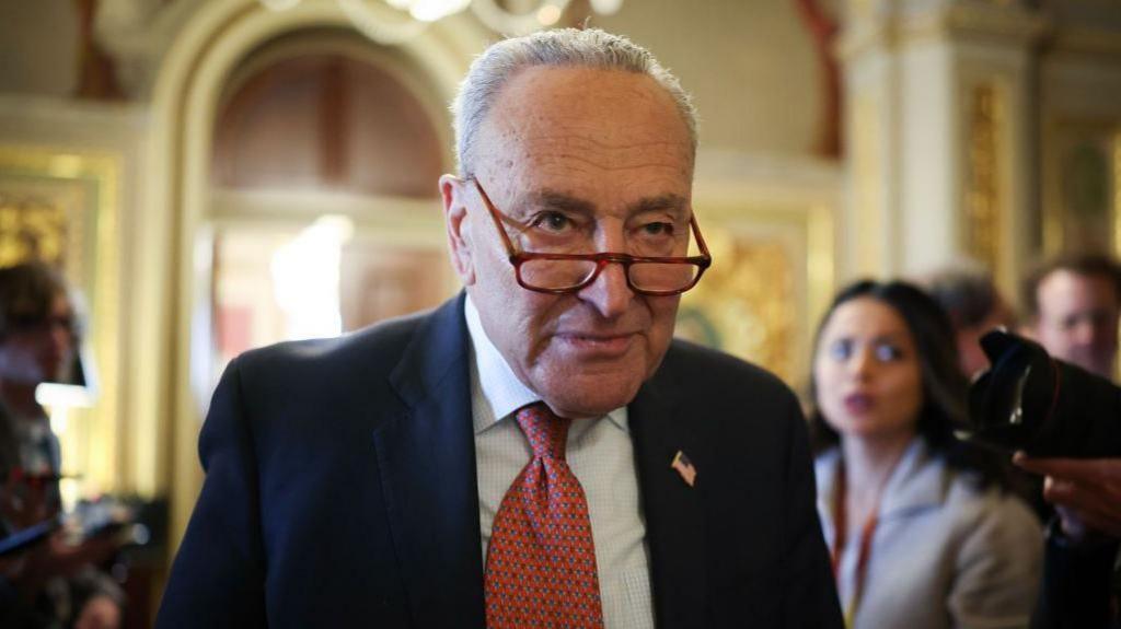 Senate Minority Leader Chuck Schumer (D-NY) leaves the Democratic caucus lunch. His glasses are perched low on his nose and his expression is neutral. He wears a blue suit and red tie, and there are reporters in the background.