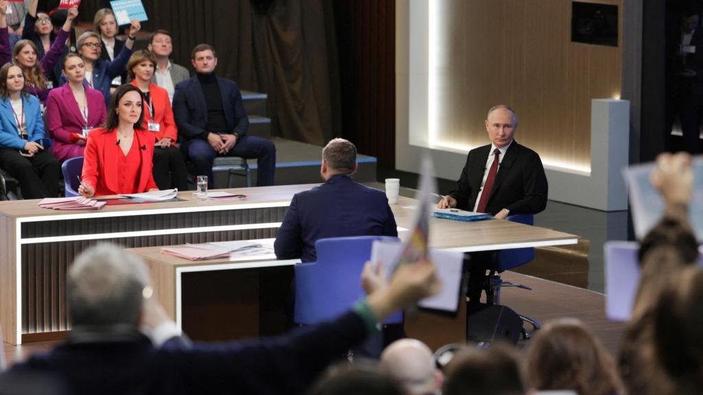 Putin sits at a desk as people in the audience holdup signs to ask a question