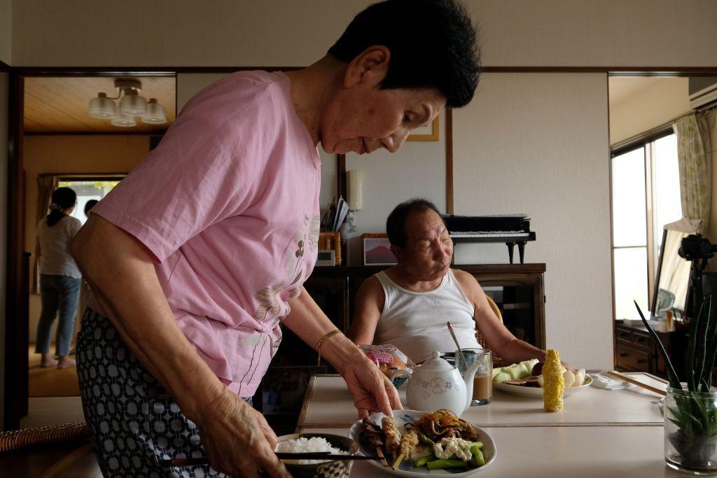 A woman sets plates of food onto a dining room table where a man in a singlet is sitting