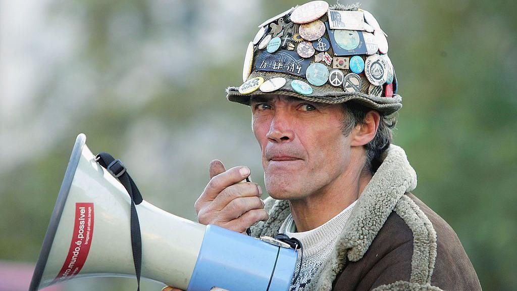 Brian Haw holds a loudspeaker in Parliament Square on 4 November 2004. He wears a had covered in pin badges and a brown coat. 