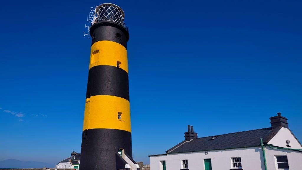 St. John's Point Lighthouse. Its strikingly tall tower is marked with vibrant bands of yellow and black. These vivid colours, which distinguish it from other lighthouses, are known as its daymark. To the right of the lighthouse is a white building with a black roof.