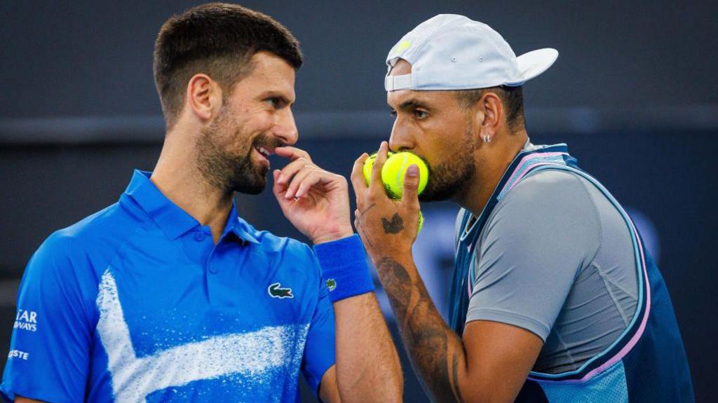 Novak Djokovic and Nick Kyrgios confrerring on court, holding their hands up to their mouths