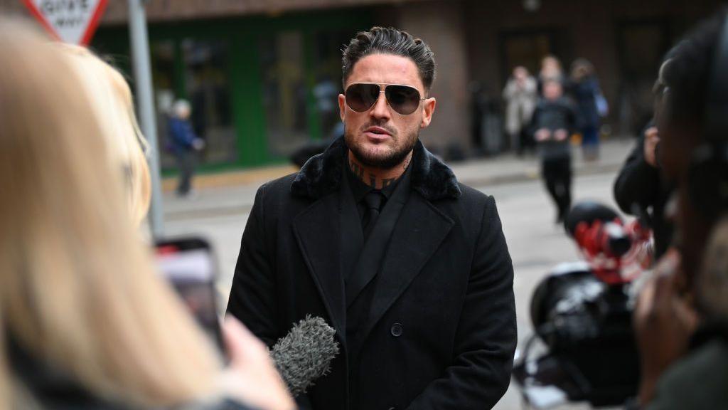 Stephen Bear with dark hair and neck tattoos, wearing sunglasses and a black coat over a black suit and tie. He is standing in the street and appears to be in the middle of speaking, with someone holding a microphone out to him.