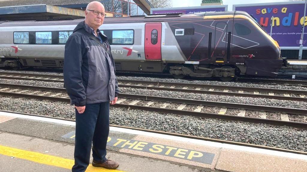 Railfuture spokesperson Dave Richardson at Oxford Train Station. There is a CrossCountry train on the track behind him. It's a cloudy day.