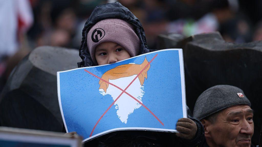 People, including a boy holding a placard that shows Greenland with the orange hair of U.S. President Donald Trump and Xed out, gather to march in protest against U.S. President Donald Trump and his announced intent to acquire Greenland on January 17, 2026 in Nuuk, Greenland